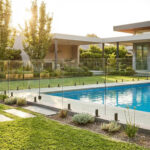 Piscine rectangulaire avec clôture de sécurité en verre, bordée d'un jardin verdoyant et d'une maison moderne baignée de soleil.